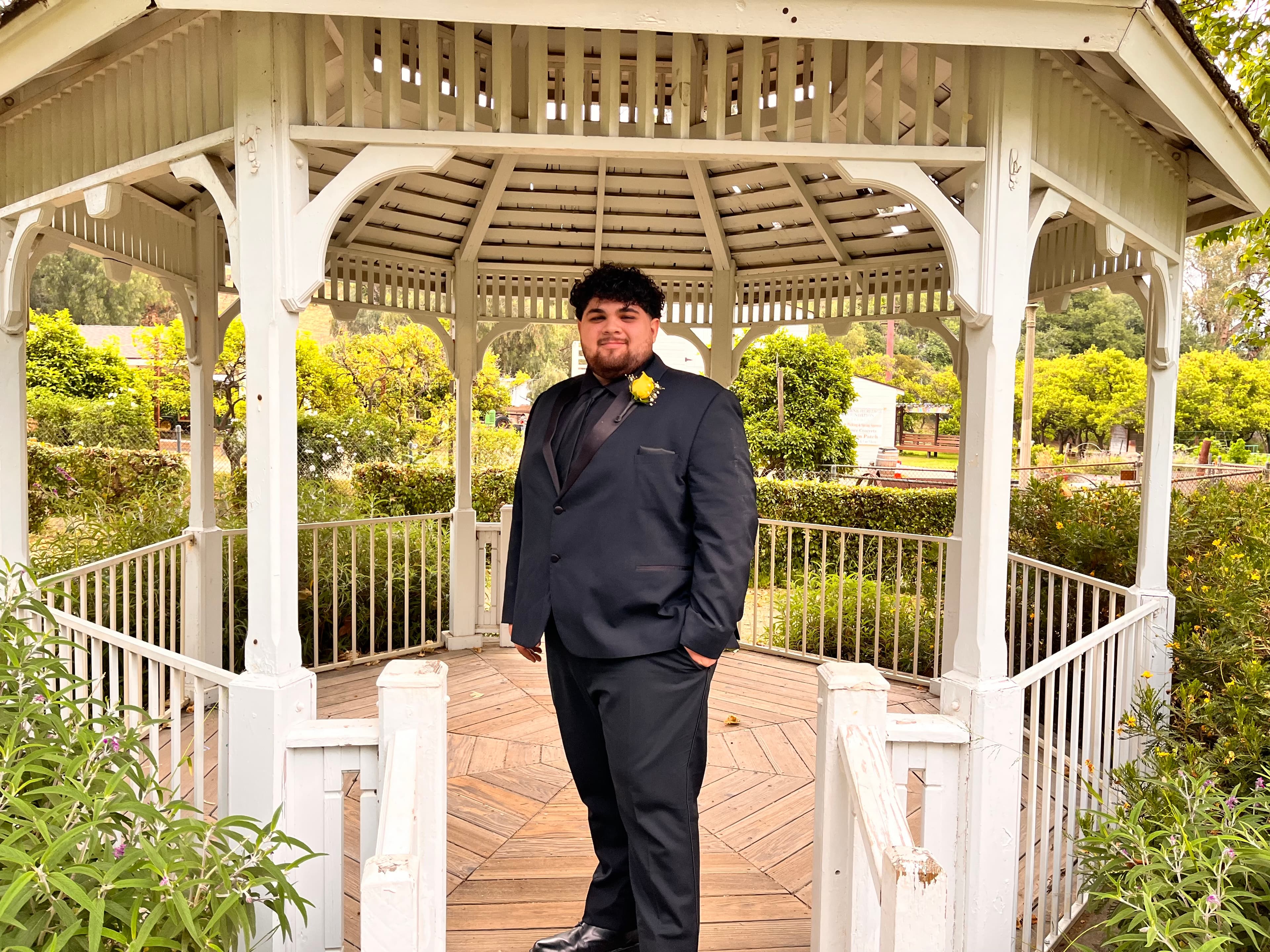 Smiling man in a dark suit and yellow boutonniere posing inside a white wooden gazebo.